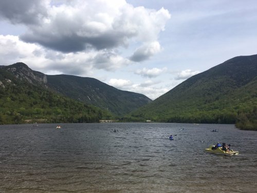 A serene lake surrounded by mountains, with people kayaking and cloudy skies above.