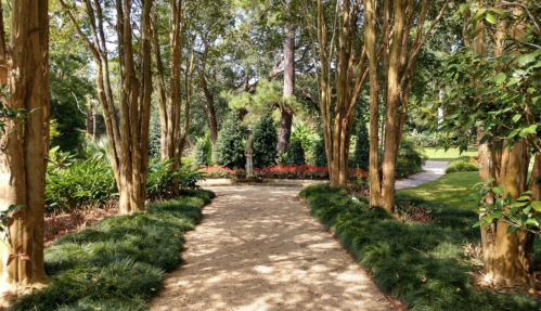 A serene garden path lined with trees, leading to a statue surrounded by vibrant flowers and lush greenery.