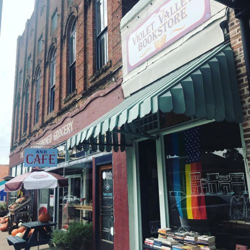 A quaint street scene featuring a bookstore with a rainbow flag, a café, and autumn decorations outside.