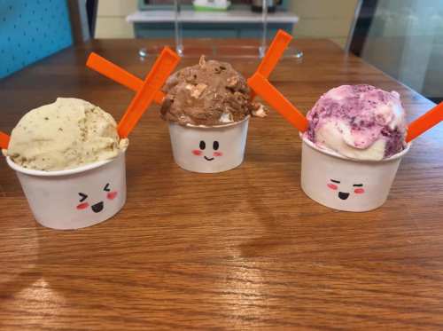 Three cups of ice cream with smiling faces: one green tea, one chocolate, and one berry, each with orange spoons.