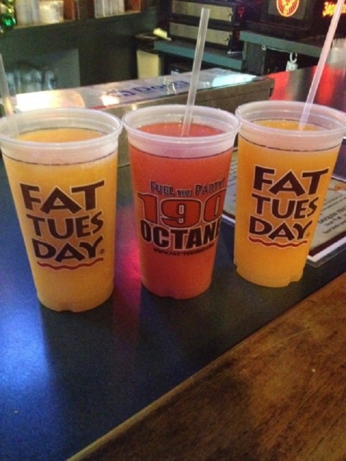 Three colorful drinks in clear cups on a bar counter, labeled "Fat Tuesday" and "Fuel the Party."
