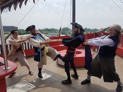 Four people dressed as pirates playfully tug on a rope aboard a ship, with a bright sky in the background.