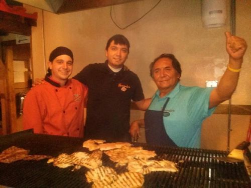 Three chefs pose together in a kitchen, smiling, with grilled meats on a large grill in front of them.