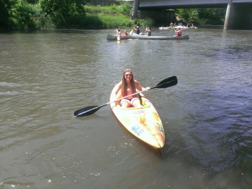 A person in a kayak paddles on a river, with other kayakers visible in the background under a bridge.