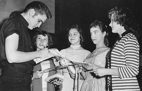 Elvis Presley signs autographs for four young women in a vintage black-and-white photo.