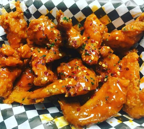 A close-up of spicy chicken wings coated in a glossy orange sauce, garnished with herbs and red pepper flakes.