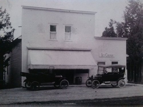 Historic black-and-white photo of an ice cream shop with two vintage cars parked outside.