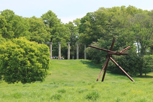 A grassy landscape featuring a large wooden sculpture and stone columns surrounded by trees.
