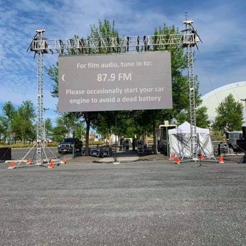 Outdoor setup for a film screening, featuring a large screen with audio instructions and a reminder to start car engines.