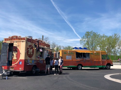 Two food trucks parked in a sunny area, with people ordering food and trees in the background.