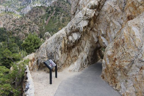 A rocky tunnel with a pathway leading through it, surrounded by lush greenery and mountains in the background.