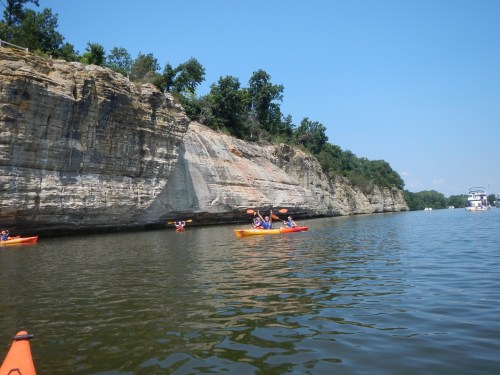 Two kayakers paddle on a calm river near a rocky cliff under a clear blue sky. A boat is visible in the background.