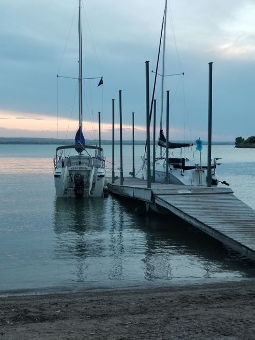 Two sailboats docked at a wooden pier on a calm lake during twilight, with a cloudy sky in the background.