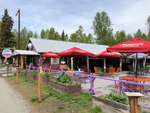 A rustic building with outdoor seating, colorful chairs, and red umbrellas, surrounded by greenery on a sunny day.