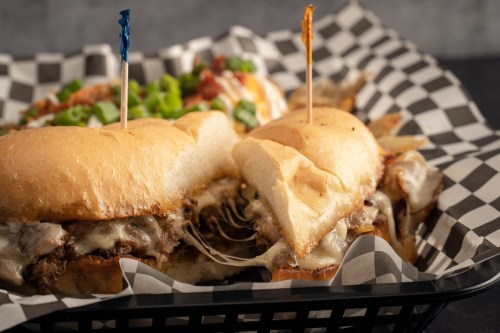 Two cheesy sandwiches with toppings, served in a black and white checkered basket, alongside a side of fries.