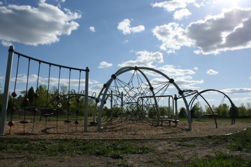 A playground with climbing structures and a net, set against a blue sky with fluffy clouds and green trees in the background.
