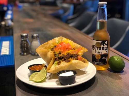 A taco salad in a crispy shell, topped with beef, cheese, tomatoes, and served with salsa, sour cream, a lime, and a beer.