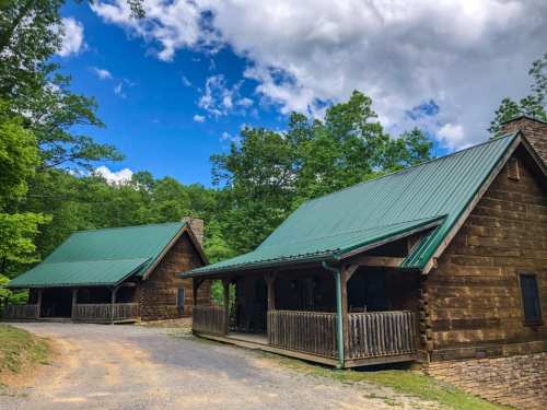 Two rustic cabins with green metal roofs surrounded by lush trees under a partly cloudy blue sky.