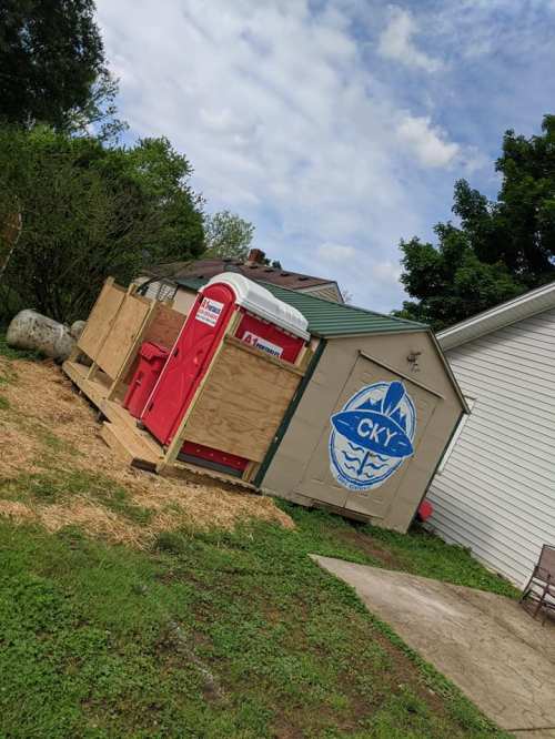 A portable restroom next to a shed, surrounded by wooden fencing and straw, under a cloudy sky.