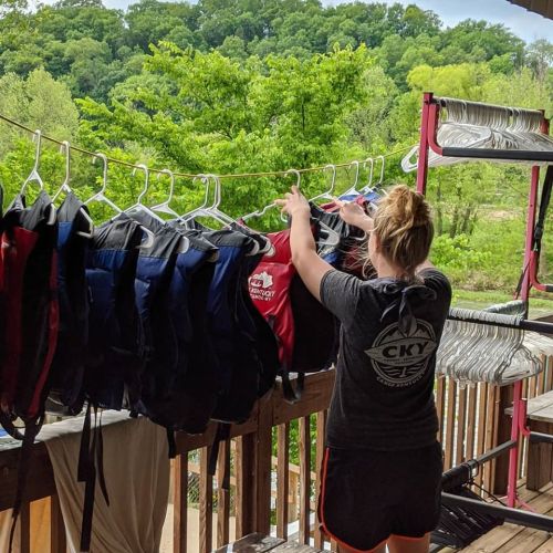 A person hangs life jackets on a rack outdoors, surrounded by greenery and a wooden structure.