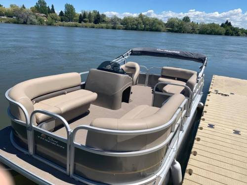 A spacious, beige pontoon boat docked by a calm river with green trees in the background under a clear blue sky.