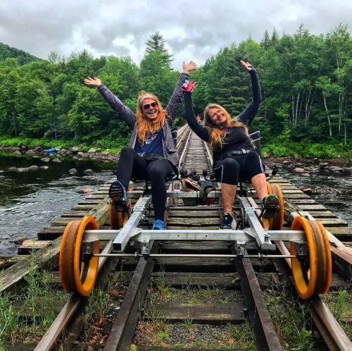 Two women joyfully pose on a rail bike on a wooden bridge, surrounded by lush greenery and a river.