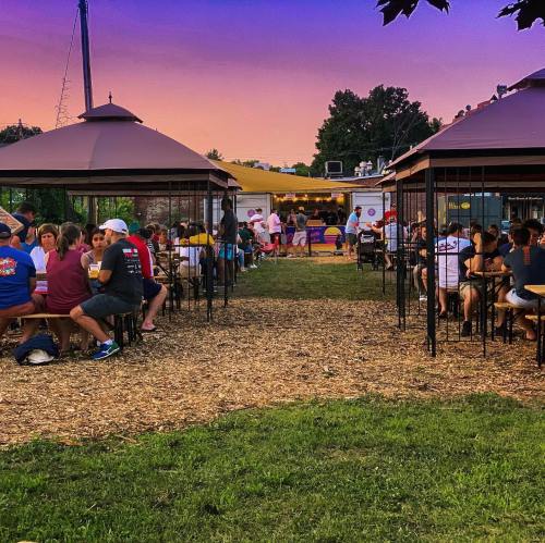 Outdoor dining area with people seated at tables under gazebos, enjoying food and drinks at sunset.