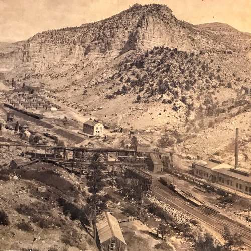 A sepia-toned landscape featuring a mining town with buildings, tracks, and mountains in the background.