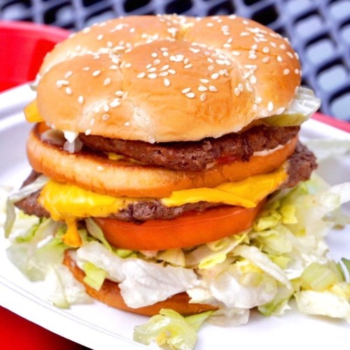 A towering burger with two patties, melted cheese, lettuce, tomato, and sesame seed bun, served on a white plate.