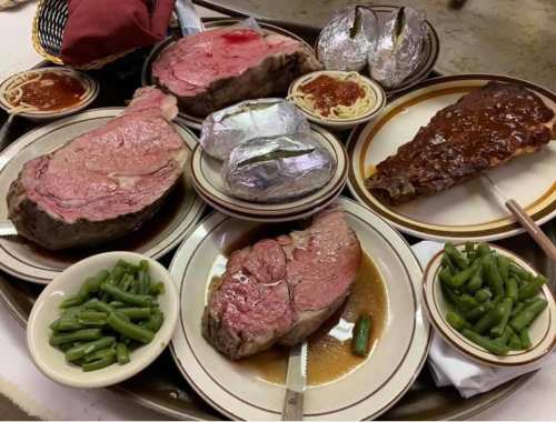 A large platter featuring prime rib, ribs, baked potatoes, green beans, and fish, served with sauce and spaghetti.