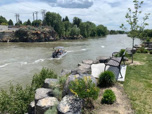 A river flows past a park with a bench, rocks, and greenery, while a boat travels on the water under a cloudy sky.