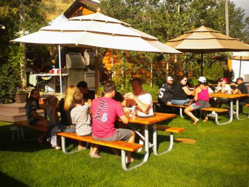 A group of people enjoying food and drinks at picnic tables under umbrellas in a green outdoor setting.