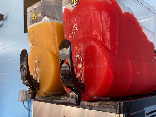 Two beverage dispensers: one filled with yellow juice and the other with red slush, both on a stainless steel base.