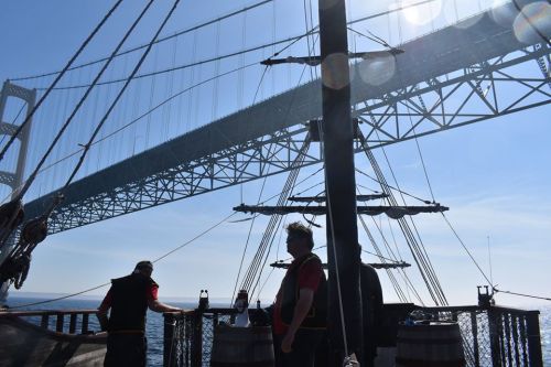 Two crew members on a ship under a large bridge, with sunlight reflecting off the water and the structure above.