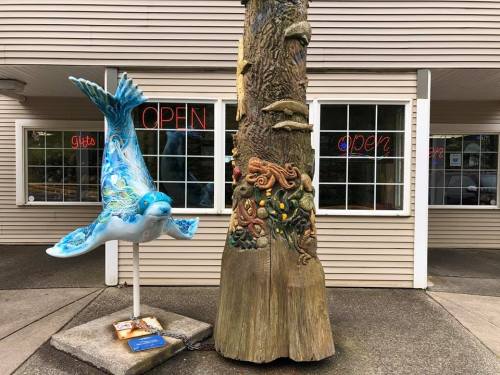 A colorful bird sculpture and a carved tree trunk stand outside a building with "Open" signs in the windows.