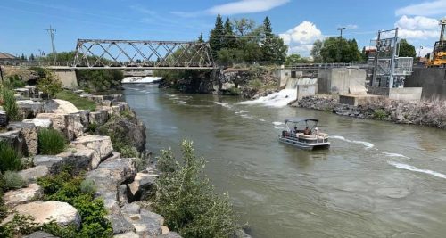 A boat navigates a river near a rocky shore and a bridge, with greenery and a dam in the background under a blue sky.