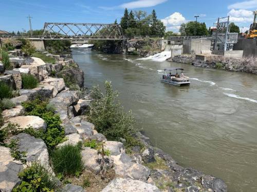 A boat navigates a river near a rocky shore and a bridge, with greenery and a dam in the background under a blue sky.