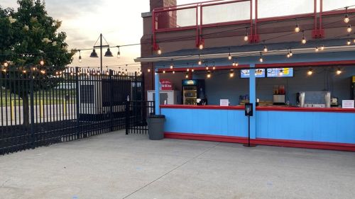 A colorful outdoor concession stand with string lights, a fence, and a trash can nearby, set against a sunset sky.
