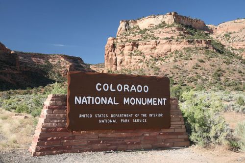 Sign for Colorado National Monument, featuring rugged rock formations and desert vegetation in the background.