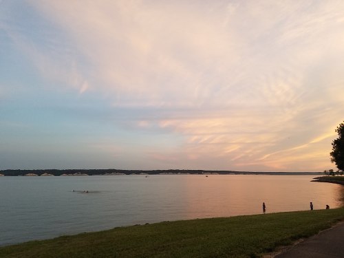 A serene lakeside view at sunset, with soft clouds and a few people enjoying the shoreline.