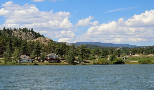 A serene lake scene with houses along the shore, surrounded by trees and mountains under a blue sky with fluffy clouds.