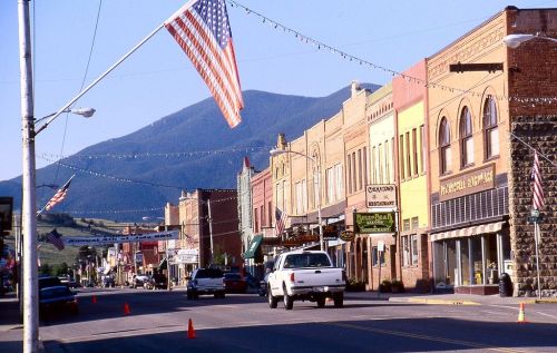 A small town street lined with colorful buildings, American flags, and mountains in the background on a clear day.