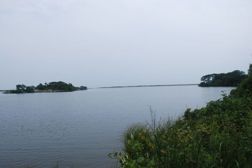 A calm, overcast lake scene with distant islands and lush greenery along the shoreline.