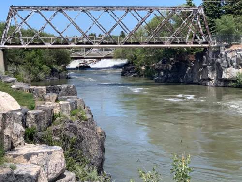 A weathered metal bridge spans a river, surrounded by rocky banks and lush greenery under a clear blue sky.