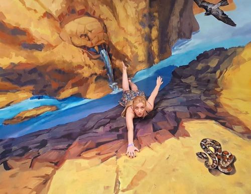 A woman poses on a 3D mural, appearing to hang over a rocky cliff with a waterfall and a snake nearby.
