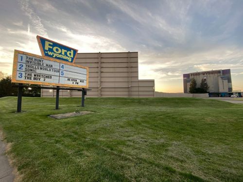 A marquee outside Ford Wyoming theater displays movie titles with a large building in the background under a colorful sky.