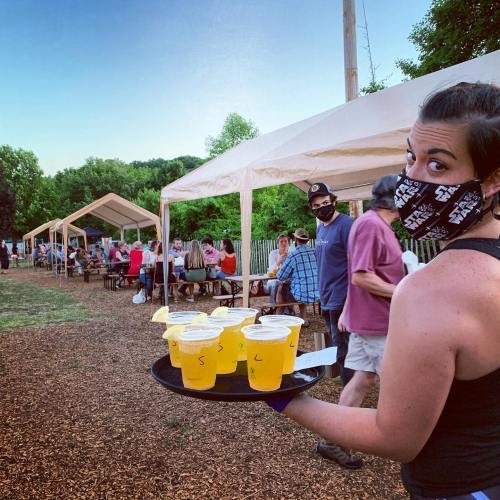 A server in a mask carries a tray of drinks at an outdoor gathering with people seated at tables in the background.