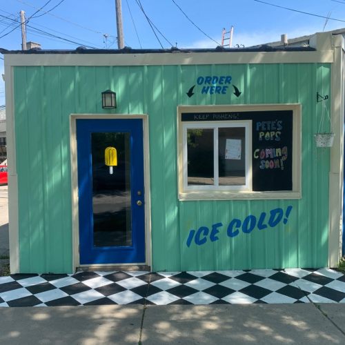 A colorful ice pop shop exterior with a blue door, "Order Here" sign, and "Ice Cold!" painted on the wall.