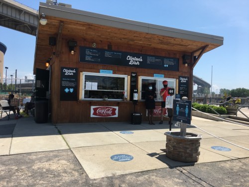 A wooden food stand named "Clinton's Dish" with a menu, outdoor seating, and people ordering in a sunny setting.