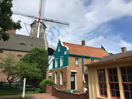A traditional windmill beside colorful brick houses under a partly cloudy sky. Green trees add to the scenic view.